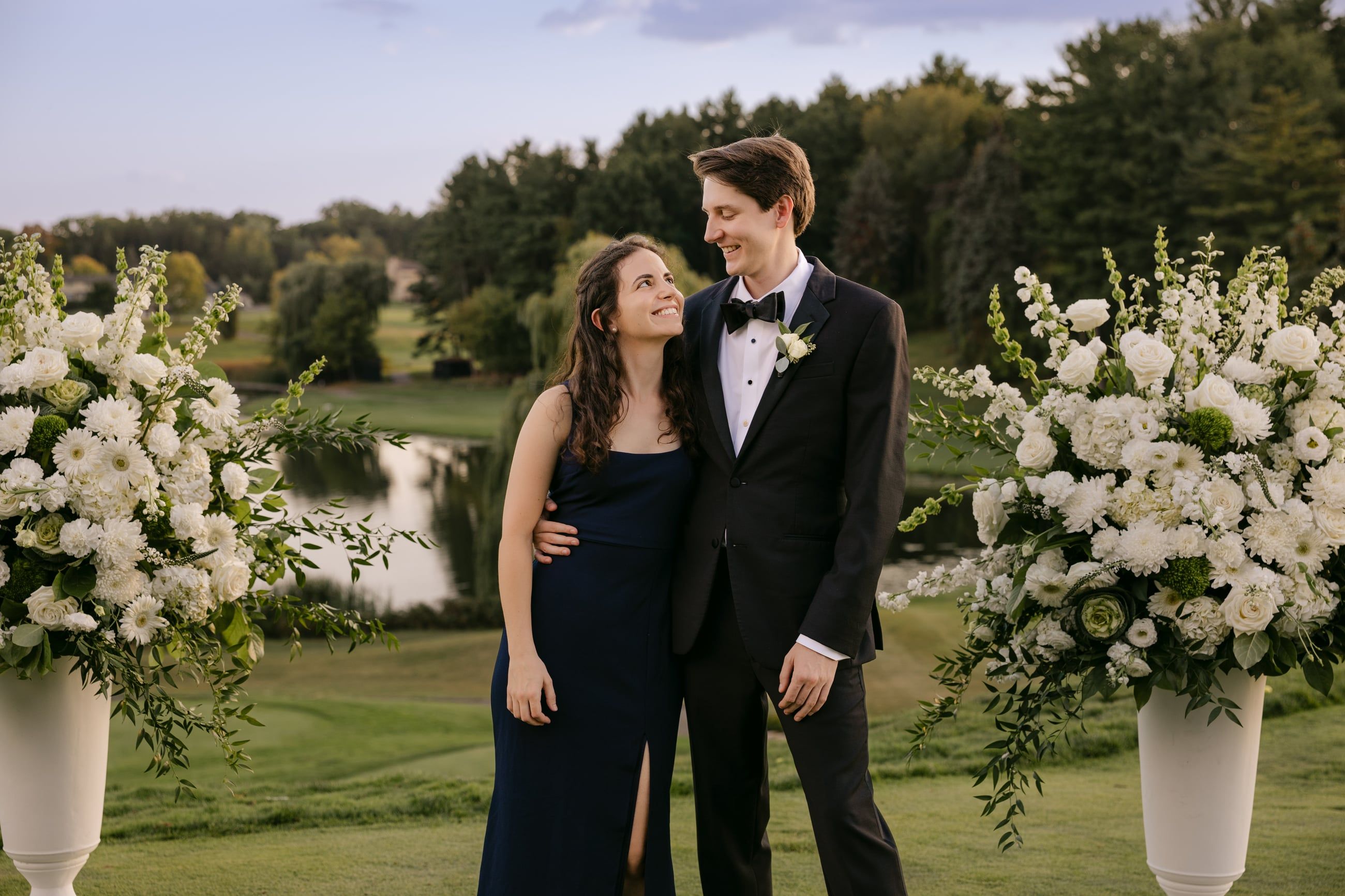 Katherine and Johnny arm in arm between two urns of white flowers at a garden wedding