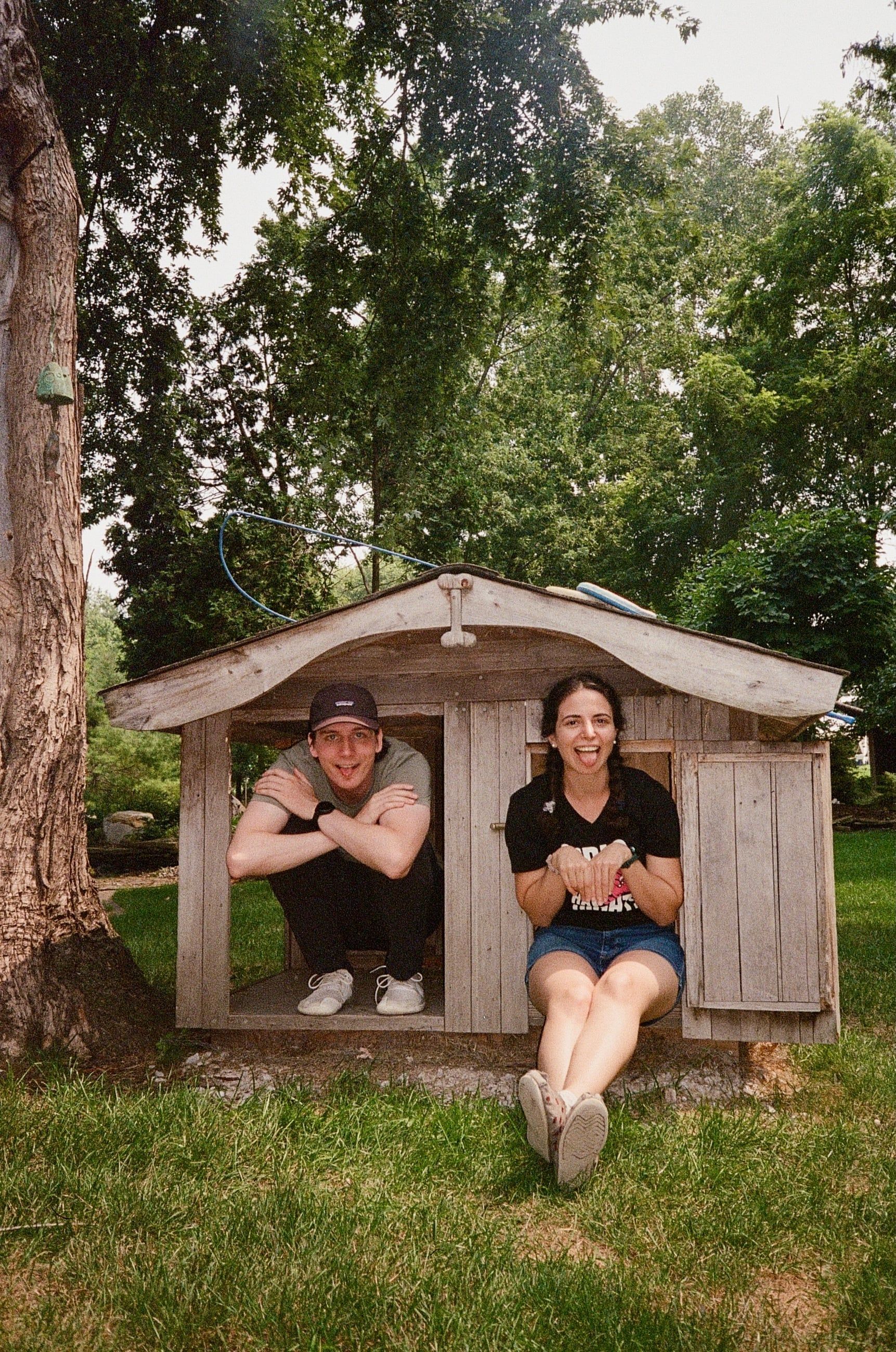 Katherine and Johnny sitting in the doorway of a weathered wooden playhouse under a big tree